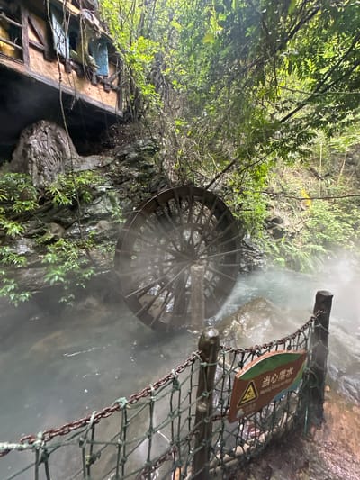 The image shows a water wheel in a stream, with a wooden structure built into a rocky cliff face behind it. The area is lush with vegetation, and a mist or steam rises from the water. A signpost with Chinese characters and an English warning about falling into the water is attached to a chain-link fence in the foreground. The water wheel appears to be made of wood and is partially submerged in the water, with its spokes visible. The wooden structure on the cliff appears to be an unusual building with windows and what looks like a balcony. This scene is located in the city of Qingyuan City, China, specifically in the area of the Huangteng Gorge, which is known for its hot springs and natural scenery. There are no people visible in the image.