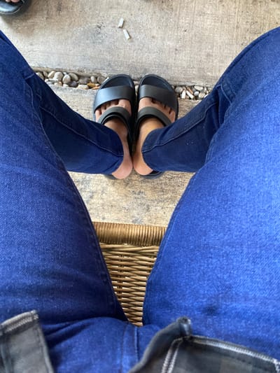 A first-person perspective shot shows a person sitting, with their legs crossed. The person is wearing dark blue denim jeans and black slide sandals. The background consists of a concrete walkway with small pebbles and two cigarette butts, and a woven cane chair. The image appears to have been taken outdoors in a location that might be found in Aluepiet, Indonesia, though no specific landmarks or distinctive city features are visible.
