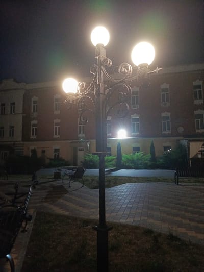 This image depicts a lamppost with three illuminated globes in the foreground, casting a warm light on the surrounding area. Behind the lamppost, a multi-story brick building with numerous windows is visible. The ground in front of the building is paved with interlocking bricks, and there are benches and greenery. The scene appears to be a public space or park, likely in the city of Biaroza, Belarus, as suggested by the context. The time is night, indicated by the darkness of the sky and the artificial lighting. There are no people or animals present in the image, and no actions or interactions are occurring.