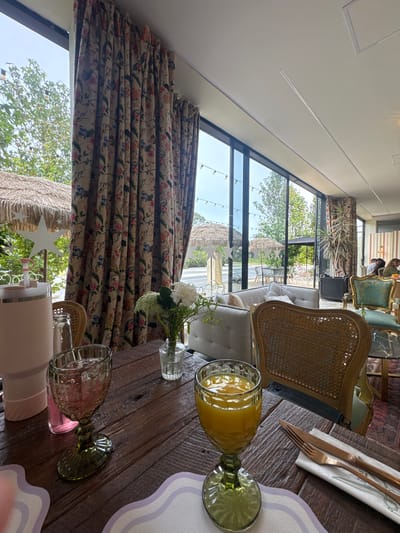This media file depicts a close-up view of a wooden table set for a meal or drinks, with a backdrop of an indoor seating area featuring large windows and outdoor scenery. The table is made of rustic, wide wooden planks. On the table, there are two decorative glassware items. One glass, with a textured green tint, is filled with an orange liquid, likely juice. Beside it, a white napkin is folded, holding a set of rose gold-colored cutlery – a fork and a knife. Another green-tinted glass, this one appearing empty or containing ice and a pink liquid, is positioned closer to the foreground. A small vase containing white flowers and greenery sits near the orange juice glass.

Beyond the table, through large windows, a lush green outdoor environment is visible. Trees and what appear to be thatched-roof structures suggest a garden or patio area. String lights are strung across the outdoor space. Inside, the seating area is furnished with upholstered sofas and chairs, including a distinctive rattan-backed chair in the mid-ground. The interior design includes floral patterned curtains that cover some of the windows. The lighting suggests it is daytime, likely late morning or afternoon, with natural light streaming in. The overall impression is a relaxed and possibly upscale dining or lounge setting in a location that combines indoor comfort with a connection to nature. The image does not contain any visible text.