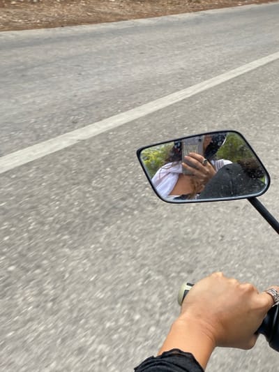 The image presents a first-person perspective from a moving vehicle, likely a scooter or motorcycle, on a paved road in Aegina, Greece. In the bottom right foreground, a hand with light skin, wearing a silver-toned, intricately designed ring on the fourth finger, grips a black and light-colored handlebar. The cuff of a black, ruffled sleeve is visible on the wrist. A black rectangular rearview mirror dominates the mid-right portion of the frame. The mirror's reflection captures a person, identified as the driver, holding a grey smartphone to take the photograph. This person exhibits long, dark, curly hair, wears a white garment over a darker top, and has a partially visible black helmet. A ring is also visible on a finger of the hand holding the phone. Lush green foliage is reflected behind the person. The environment consists of a grey asphalt road with a faded white lane line, indicating forward motion. A sparse, dry embankment is visible along the left side of the road.