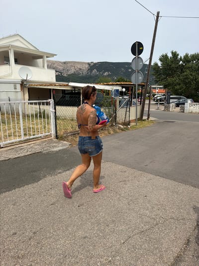 A woman with tattoos on her back walks on a paved road in Općina Baška, Croatia. She is wearing a blue bikini top, denim shorts, and pink flip-flops. She is carrying a large blue water jug and a pink object in her arms. To her left is a white fence and a building with a satellite dish. In the background, there are trees, cars, and a hilly landscape under a cloudy sky. A signpost with three circular signs is visible to her right.