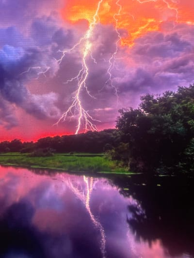 The image depicts a dramatic stormy landscape located in the city of Zelenyi Hai, Ukraine. A prominent, intensely bright white-yellow lightning bolt descends from the upper right quadrant of the sky, striking towards the horizon on the left. The sky exhibits a vivid and saturated color palette: fiery orange and red hues are concentrated towards the upper right and the distant horizon, gradually transitioning into deep purples, blues, and grays within dense cloud formations across the central and left portions. A dark, dense tree line forms the horizon, silhouetted against the bright red sky. Below the tree line, a strip of vibrant green land is visible. The foreground is dominated by a body of water, likely a river or lake, which mirrors the dramatic sky in shades of purple and pink, clearly reflecting the shimmering lightning bolt. No human subjects, animals, or man-made structures are overtly visible within the frame.