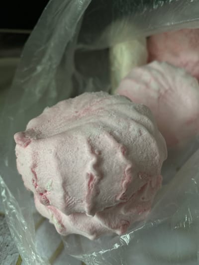 The image shows a close-up of several pink, fluffy confections, likely marshmallows or meringues, contained within a clear plastic bag. The confections are large and appear to be dusted with a fine white powder, possibly powdered sugar. The texture is soft and airy, with visible ripples and folds on their surfaces.
One confection is prominently featured in the foreground, taking up a significant portion of the frame. Its pink color is a light pastel shade, with some areas appearing slightly lighter or whiter where the dusting is more concentrated. There are hints of deeper pink or red within the confection, suggesting it might be flavored or dyed. The plastic bag's transparency allows for a slightly diffused view of the items inside and the surrounding environment, though the background is largely out of focus.
Behind the main confection, at least one other similar item is visible, slightly blurred and positioned further back in the bag. A small portion of what appears to be a pale, creamy-colored object or layer is visible beneath the pink confections, hinting at a possible base or another component of the treat.
The lighting is soft and diffused, suggesting an indoor setting or an overcast day. There are no discernible people or any text visible in the image. The focus is entirely on the texture and appearance of the sweets. Given the location context of Shostka, Ukraine, these confections could be a local delicacy or a product from a bakery in the region. The overall impression is one of sweet indulgence and delicate texture.