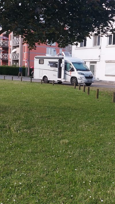 A white campervan is parked on a street next to a grassy area with scattered white flowers. A person in a dark blue shirt and grey pants is walking away from the campervan on the sidewalk. In the background, there are multi-story buildings with balconies and a tree with dark green leaves overhangs the scene. The license plate of the campervan is visible and reads "SW 93104". The location is in the city of Namur, Belgium.
