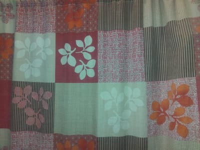 A close-up, overhead shot shows a fabric curtain composed of a patchwork of square and rectangular sections. The fabric is creased and appears to be hanging. The sections feature various patterns and colors: some have a red background with white leaf designs, others have an orange leaf design on a white patterned background, and there are also sections with thin vertical stripes in dark gray and white, and sections with a light green checkered pattern. The overall impression is of a decorative fabric with a variety of textile designs. The image is a flat lay of the fabric, not depicting any environment or specific location within Haveli Jhangranwali, Pakistan.