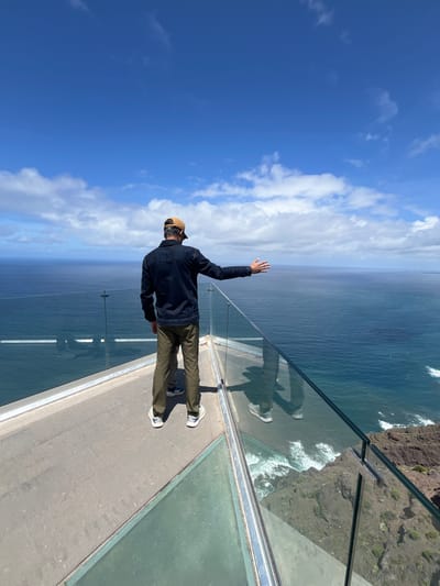 A person is standing on the Mirador del Balcón viewpoint in La Aldea de San Nicolás, Spain. The viewpoint has a glass floor and railing offering a view of the ocean. The person is wearing a dark jacket, tan pants, a tan baseball cap, and white shoes. Their left arm is raised in a wave. The sky is blue with some clouds. The cliffs and waves crashing against them are visible beneath the glass floor. The image is taken from a high angle, looking down and out toward the horizon.