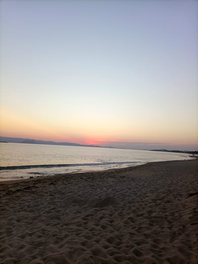 This is a wide-angle, landscape photograph of a beach at sunset in Naxos, Greece. The scene is dominated by the sandy beach in the foreground, with gentle waves lapping the shore in the middle ground. The sea stretches out to the horizon, where it meets a sky painted with the soft hues of sunset, transitioning from pale blue at the top to warm pinks and oranges near the horizon. Distant hills or mountains form a silhouette against the sky. A few small figures of people can be seen walking along the distant shoreline, indicating human presence but not as a central focus. The overall atmosphere is serene and tranquil.