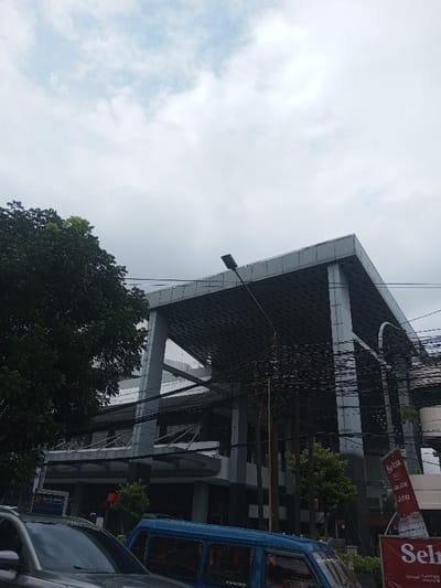 The image captures an urban street scene dominated by a modern building under an overcast sky. The sky, occupying the top portion of the frame, is light blue with diffuse white and grey clouds, indicating a cloudy or overcast day with soft, diffused lighting.
A large, contemporary building is a prominent feature, extending from the middle-left to the right side of the image. It features a striking architectural design with multiple stories and a distinctive, large cantilevered or overhanging roof structure. The building's exterior appears to be clad in grey or silver panels, giving it a sleek, modern look. The underside of the overhang has a grid-like or textured pattern. Several sturdy vertical pillars support the upper sections of the building. On the lower floors, windows are visible, and towards the left, a smaller entrance with a canopy is discernible.
On the left side of the image, a large, dark green leafy tree stands tall, its dense foliage partially obscuring the building and reaching towards the sky. Smaller green vegetation is also visible at the ground level near the building.
Numerous black power lines or utility cables crisscross the mid-ground of the image, running in front of the building and the sky, creating a busy network. These lines are supported by several utility poles, visible particularly on the right side. A black street light fixture on a pole extends from the mid-right foreground towards the upper center, positioned in front of the building's overhang.
At the bottom of the image, parts of several vehicles are visible, suggesting a street or parking area. On the far left, the hood and windshield of a silver or dark-colored car are present. In the center-bottom, the side and windows of a blue van, possibly an older model, are clearly seen. Faint text like "ESPRES" or similar branding can be seen on the side of the blue van. On the far right, a partially obscured red banner or sign is visible, featuring white text that includes "Sera", "ck", and "jda", with a clearer "Seira" below it, likely a brand name. Another blue sign, mostly hidden by the silver car on the left, shows partial text "63 PURI".
The overall impression is a bustling urban environment with a mix of modern architecture, natural elements, and infrastructure.