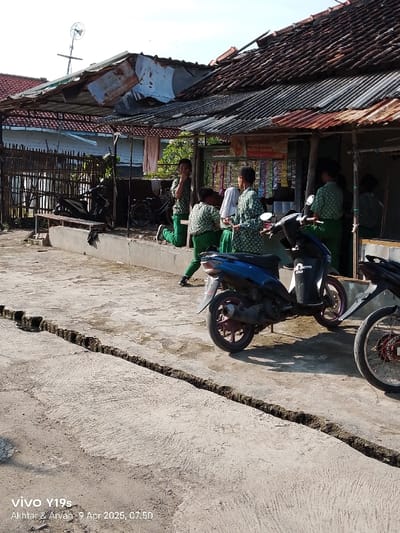 The scene is in front of a small shop with a corrugated metal roof in Kecamatan Gegesik, Indonesia (latitude: -6.536, longitude: 108.43). Five young men, dressed in green shirts and pants, are standing around a blue motorcycle with a black seat and a black fender. Two of the men are leaning against the motorcycle. One man is walking toward the motorcycle while another is sitting on the seat. The fifth man is walking away from the motorcycle toward the shop. There is a blue and white motorcycle behind the blue motorcycle with a man seated on it. The shop is located on a street with a cracked sidewalk. The shop is shaded by a roof that is partially broken. There is a wooden fence and a small patch of grass on the left side of the image. The shop has a large sign on the wall with writing that is not visible.