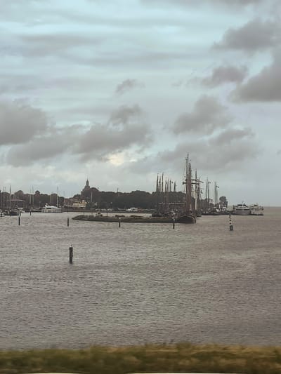 The image depicts a harbor in Enkhuizen, Netherlands, under a cloudy sky. Several sailboats and a ferry are visible in the water, along with buoys and a distant cityscape. The water is calm, and the overall atmosphere is subdued due to the overcast weather. The harbor appears to be a popular spot for sailing enthusiasts, with a variety of vessels docked and moored. The cityscape in the background suggests a historical and charming Dutch town.