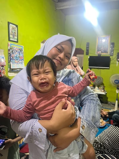 In a room with a bright lime green wall, a young woman wearing a light blue denim jacket over a striped blue and white shirt, and a white hijab, is holding a crying baby. The baby, wearing a pink long-sleeved shirt, is being held in a cradling position, with one arm around its back and the other supporting its legs. The baby's mouth is open in a wail, its eyes are closed, and its arms are outstretched. The young woman is smiling and looking towards the camera, her left arm also cradling the baby. She is holding a small red toy car in her right hand. In the background, a framed picture hangs on the wall, along with two speakers. A television is visible above a black surface, and a woman wearing a pink hijab is seated behind the young woman. Further in the background, a pedestal fan is visible, and the lower right corner shows a person wearing a striped black and white top. The overall impression is of a candid indoor moment. This scene is located in Wedarijaksa, Indonesia.