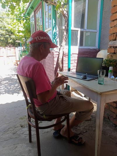 A man wearing a red cap with "TÜRKİYE" written on it and a pink t-shirt sits at a white table outdoors, looking at a laptop. He is wearing khaki shorts and dark sandals. On the table are the laptop, a computer mouse, a glass of water, sunglasses, a bottle of insect repellent, and a potted plant. The scene appears to be set in a yard, with a brick wall to the right and a green wooden building with windows behind the man. Trees with green leaves provide shade, suggesting it is daytime and likely warm weather. The ground is concrete with some cracks. The man's hands are visible, and he appears engaged with his laptop. The overall impression is of a casual, possibly domestic setting in Salhany, Ukraine.