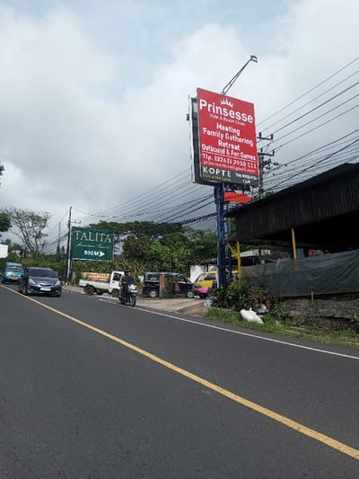 The image displays a roadside scene under a cloudy sky. The location is identified as Saing Ta Mawt Lay, Myanmar; however, prominent signage for "Prinsesse Hotel & Resort Ciloto" and "TALITA Mountain Resort" suggests a location in Ciloto, Indonesia. The asphalt road, marked with a solid yellow center line, extends from the lower-left to the upper-right.
**Vehicles:** A dark-colored motorcycle, ridden by an individual wearing a dark helmet and jacket, is in motion in the foreground, moving away from the viewer. Behind it, a dark grey minivan (possibly a Honda Mobilio) and a light blue minivan are also in motion. On the right shoulder, a white pickup truck with "KONTRAK" visible on its side is parked, followed by a black minivan, a yellow minivan, and a partially obscured pink minivan.
**Structures and Objects:** On the right side of the road, a large red billboard for "Prinsesse Hotel & Resort Ciloto" advertises "Meeting, Family Gathering, Retreat, Outbound & Fun Games" along with contact information and mentions "KOPTE KOPI & TEH TARIK" and "THE WEEKEND CAFE." This billboard is mounted on a blue metal pole structure, with an overhead arm. Numerous black electrical wires crisscross the sky, supported by utility poles alongside the road. A low-slung building with a dark corrugated metal roof and a black tarp covering part of its facade stands on the far right. Below it, a concrete wall and patches of grass with white plastic bags are visible. On the left side, behind the moving vehicles, a green sign reads "TALITA Mountain Resort 800 M" with an arrow.
**Environment:** The scene is characterized by dense green foliage and trees lining both sides of the road, with distant hills or mountains visible through the overcast sky in the background. The overall impression is a semi-urban or rural area with roadside businesses and significant natural elements.