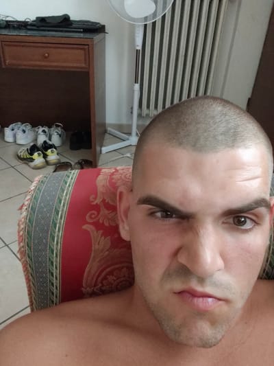 The image shows a man with a buzzed haircut sitting on a red and gold patterned cushion. He is looking at the camera with a stern expression. In the background, there is a wooden dresser with several pairs of shoes on the floor beneath it, as well as a white oscillating fan and a radiator. The man's skin appears tanned and there are no visible injuries. The scene suggests an indoor setting, possibly a home. The metadata indicates the image was taken in Guarene, Italy.