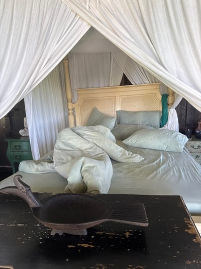 An interior shot depicts a bedroom in Tibubiyu, Indonesia, viewed from a low angle. In the foreground, a dark, distressed wooden table features a dark brown, carved wooden bowl resembling a stylized duck or bird, with an elongated neck and textured tail fin, positioned centrally. The midground is dominated by a large four-poster bed with a light-colored wooden headboard featuring two textured, rectangular panels. The bed is covered in light teal or pale green bedding, including a fitted sheet, several pillows, and a rumpled duvet, suggesting recent use. White translucent canopy drapes are pulled back from the bedposts, framing the scene. To the left of the bed, a dark green wooden nightstand with two drawers and brass pull handles supports a light-colored, possibly white, statuette or decorative object. To the right, a dark, carved wooden screen or panel is visible, with a light-colored object situated on a distressed cabinet behind it. No individuals are present.