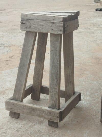 A rough-hewn wooden stool or stand stands on a sandy, concrete-like surface in Yola, Nigeria. The object, made from weathered planks and secured with nails, has a square top supported by four legs. Two of the legs are angled, while the other two are perpendicular. A horizontal bracing piece connects the bottom of the legs, forming a square base. The background is indistinct, showing a continuation of the sandy surface and a slight elevation in the ground. No people or animals are visible in the frame.