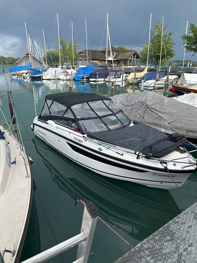 The image depicts a marina on Chiemsee, Germany, with a fleet of sailboats and motorboats docked under a cloudy sky. In the foreground, a white motorboat named "Sunrise" is prominently displayed, its canopy up and a black cover on its bow. Adjacent to it, on the left, is the hull of another boat. In the background, numerous sailboats, many covered with blue and white tarpaulins, line the dock. Traditional wooden boathouses with steep roofs are visible behind the boats, further suggesting the location's setting. The water is calm and reflects the overcast sky and the surrounding vessels. The overall scene conveys a sense of quietude, with the boats at rest in the harbor. There are no people visible in the image.