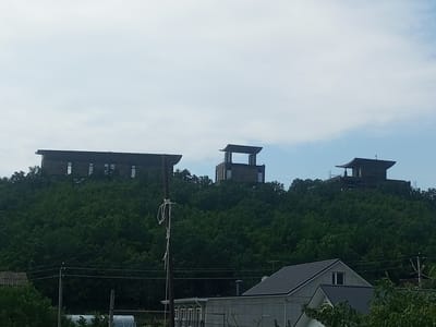 The image depicts a hilly landscape covered in dense green trees. Three modern, multi-story structures with dark, angular roofs are perched on the crests of these hills, overlooking a lower-lying area with residential buildings and utility poles. The structures appear to be constructed from stone or brick, with large windows. In the foreground, a weathered utility pole with a rope tied to it stands prominently. Power lines are visible crisscrossing the scene. The sky is overcast with a light blue hue. The location is specified as Щебетовский поселковый совет, Ukraine.