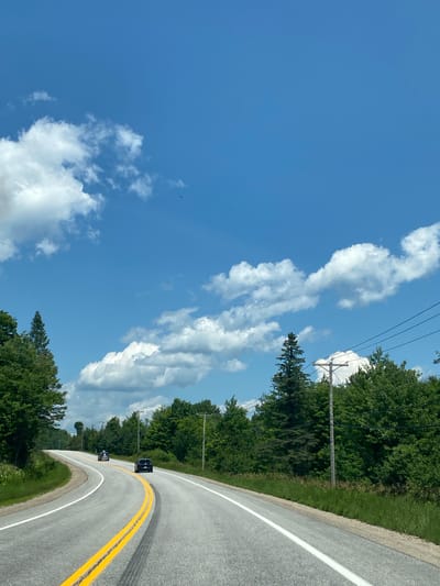 The image depicts a two-lane asphalt road curving through a rural landscape under a bright blue sky with scattered white clouds. The road is marked with double yellow lines in the center and white lines on the edges. Several dark-colored cars are visible on the road, with one in the foreground and two further ahead. The roadside is lined with lush green trees and vegetation. Telephone poles with wires are interspersed along the side of the road. There are no visible buildings or signs of a city. The scene appears to be a sunny day in a natural environment, not within the city of Ripon, Canada, as there are no urban features present.