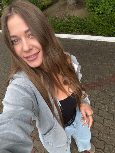 A young woman with long brown hair, wearing a gray zip-up hoodie over a black top, and light-wash denim jeans, is lying on a paved surface. Her left hand is visible, wearing a ring, and she is smiling at the camera. The background shows green vegetation near a white border, suggesting a park or landscaped area. The paving appears to be a patterned, interlocking brick or stone. The environment suggests an outdoor public space in a city, possibly Sosnovy, Russia.