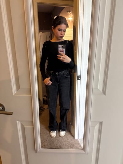 A mirror selfie reveals a woman in black long-sleeved top and black jeans. She holds a cell phone in one hand. The reflection shows a room with a light fixture. The floor is carpeted. The mirror's frame is white and hangs on a door. A doorknob is visible at the bottom.