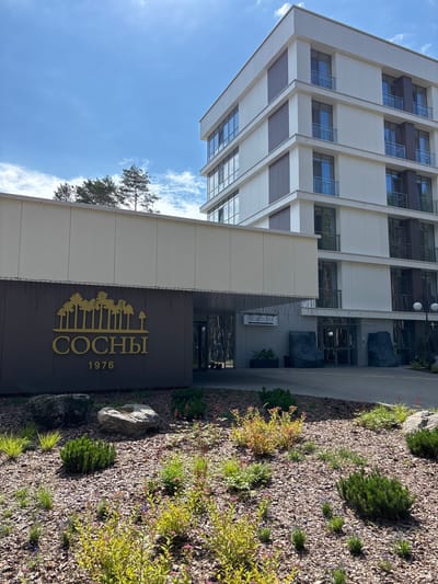 The image depicts the entrance area of a modern apartment complex named "СОСНЫ 1976" located in Мядзельскі сельскі Савет, Belarus. The building is multi-story with a light-colored facade, featuring numerous windows and balconies. In front of the building, a dark brown sign with golden lettering and an emblem of pine trees is prominently displayed. Next to the sign, a smaller sign reads "РЕЦЕПЦИЯ" (Reception). The entrance is flanked by two large, dark grey, rock-like sculptures. The foreground is landscaped with a variety of low-lying bushes, small trees, and flowering plants set in a mulch bed. The sky is blue with scattered white clouds.