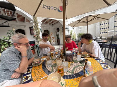 Four people are seated around a colorful mosaic table under a large umbrella. The setting appears to be an outdoor courtyard with white buildings and decorative tile work in the background. Several drinks and dishes are scattered on the table. One person is wearing a grey checkered shirt, another a white shirt. A woman is in red, and a fourth person is wearing a white shirt. They seem engaged in conversation or enjoying a meal together in a relaxed atmosphere.