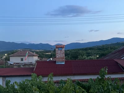 The image shows a village scene in Akandzhievo, Bulgaria, during twilight. In the foreground, a vine-covered stone wall supports a building with a red corrugated metal roof. To the left, another building with a light-colored facade and a red tiled roof has a Bulgarian flag mounted on a pole. A prominent brick chimney with a metal cap stands on a red corrugated metal roof in the mid-ground. The background features rolling hills covered in trees and distant mountains under a dusky blue sky with thin clouds. Power lines are visible stretching across the upper portion of the sky.