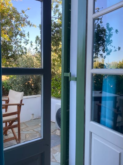 The image depicts an outdoor scene viewed from indoors, through a partially open double-leaf door or large window, located in Πλατύς Γιαλός, Greece. The left leaf of the opening features a dark gray frame with glass panes. The right leaf, partially open to the left, consists of a white frame with glass panes, reflecting an interior space that includes a blue curtain or fabric, and possibly furniture. Between the two leaves, a vertical green-painted wooden element is visible, featuring a green metal latch or hook.
Beyond the opening, an outdoor patio or terrace is visible under clear blue sky. The patio floor is paved with irregular, flat grey stones. On the left side of the patio, a wooden chair with a light-colored fabric seat and backrest is positioned. A low white wall runs along the back of the patio. To the right, against the white wall, a white outdoor air conditioning unit with a visible black fan grate is situated. Lush green foliage, consisting of trees and bushes, is prominent beyond the patio and fills the upper portion of the view, suggesting a natural or cultivated garden environment. No individuals or active subjects are present.