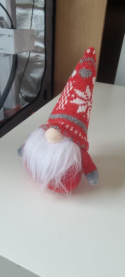A knitted gnome with a red and white snowflake patterned hat, white beard, and red body sits on a white surface. The gnome is positioned slightly to the right of the image center. In the background, to the left, is a white electrical adapter with the label "A1" and a dark grey fabric bag. To the right of the gnome, a portion of a wooden surface is visible. The scene appears to be indoors, likely on a table. The location is not identifiable as being in Naaldwijk, Netherlands.