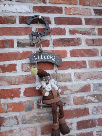 The image shows a weathered brick wall with a decorative display. A rusty horseshoe is mounted on the wall, with a padlock hanging from it. Suspended from the padlock by a rope is a small, dark-colored sign with the word "WELCOME" written in white, stylized lettering. Below the sign, a crocheted doll resembling a gnome or Santa Claus sits on a small swing. The doll is wearing a brown hat with a yellow pom-pom, a brown coat, and brown boots. Its beard is white. The doll is hanging from ropes attached to the underside of the welcome sign. The brick wall itself is constructed of red and orange bricks with white mortar, showing signs of age and wear. The overall scene appears to be an exterior decoration, possibly at the entrance to a building. The location is identified as Aalst, Belgium.