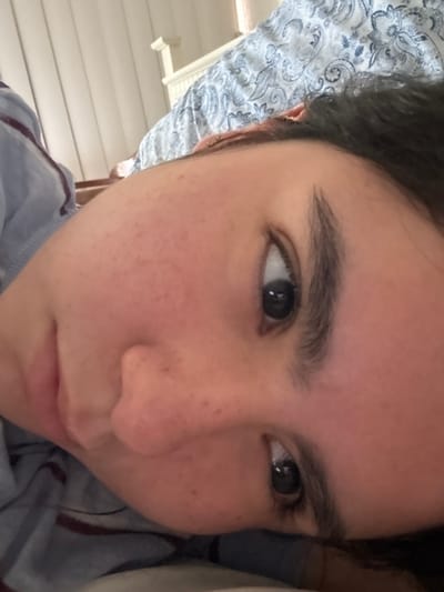 An upward-angled close-up image captures the upper portion of a person's face, rotated approximately 45 degrees counter-clockwise. The subject, likely female, has dark hair, prominent dark eyebrows, and visible eyelashes. Both dark irises are directed towards the camera. The skin appears light-toned with numerous small, dark epidermal spots, consistent with freckles or minor blemishes, across the cheeks, nose, and forehead. A slight reddish hue is visible on the cheeks. The bridge of the nose, upper lip, and the corner of the mouth are partially visible on the lower left of the frame. The upper helix of the right ear is visible, adorned with a small, circular, metallic earring. The subject is wearing a light blue garment with faint darker stripes on the left shoulder area.

The background indicates an indoor setting, likely a bedroom. A white, possibly wooden, headboard with a carved or paneled design is visible behind the subject's head. A blue and white patterned comforter or duvet with a paisley-like design is draped over the bed. To the far upper left, vertical blinds or light-colored drapes are partially visible, suggesting a window as a light source. The lighting is soft and ambient.

The image contains no identifiable landmarks, signage, or contextual elements to determine a specific location within the city of Santiago, Mexico.
