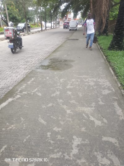 A medium-shot, eye-level, outdoor photograph captures a man walking on a sidewalk, moving from right to left. He is dressed in a white t-shirt with red and blue accents and light blue jeans. He carries a brown bag in his right hand and a reddish-brown object in his left. The man appears to be walking at a moderate pace.
The setting is a paved pathway adjacent to a road. The sidewalk is made of concrete with some patches of discoloration and water stains. To the left of the sidewalk is a brick-paved road with some parked vehicles, including a white van and a dark SUV. To the right of the sidewalk, there is a strip of green grass and several large palm trees with rough, fibrous trunks. The background shows more trees and some buildings, suggesting an urban or suburban environment. A motorcycle with a delivery box is visible on the road to the far left.
The time of day appears to be daytime, with overcast conditions suggested by the diffused light. The overall atmosphere seems casual and ordinary, with no strong emotional cues conveyed. The image is watermarked at the bottom left corner with "TECNO SPARK 10c". The location context provided is Awoyaya, Nigeria.