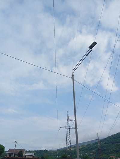 In Wazabaa, Abkhazia, the sky is a canvas of light blue with scattered white clouds. Power lines crisscross the frame, creating a complex network against the sky. A modern street lamp with a dark rectangular fixture is prominently positioned on the right, extending from a tall, gray pole. In the middle ground, a large, skeletal transmission tower stands silhouetted against the sky. To its right, another, smaller transmission tower with a red and white structure is visible. The lower portion of the image reveals a rolling, tree-covered hillside dotted with houses. In the foreground on the left, a house with a brown roof and a smaller adjacent structure are visible, along with some green foliage. The overall scene captures a semi-rural environment with significant electrical infrastructure.