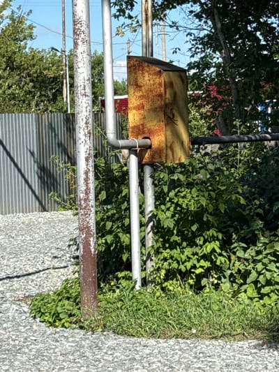 The image shows a close-up view of a rusty yellow electrical box attached to metal poles, surrounded by lush green foliage and vegetation. In the background, a corrugated metal fence is visible, along with trees and a bright blue sky. The ground in the foreground is covered in small grey gravel. The scene appears to be outdoors, possibly in a semi-rural or industrial area of Айет, Kazakhstan, given the presence of utility infrastructure and fencing. There are no people or visible signs of recent activity.