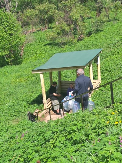 The image shows three people gathered under a wooden gazebo in a lush, green outdoor setting. One man is wearing a dark suit and appears to be handing a large water bottle to a woman who is seated on a bench. Another man is seated on the bench as well. Several large water bottles are on the bench. The gazebo has a green roof. The surrounding environment is filled with vegetation, including grass, flowering plants, and bushes. The image was taken in a natural setting. There is no indication of Avdot’ino, Russia.