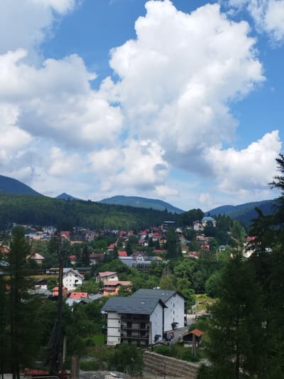 The image depicts a panoramic view of the town of Bușteni, Romania, nestled at the foot of the Bucegi Mountains. The scene is dominated by lush green forests covering the mountainsides, with scattered houses and buildings forming the town below. The sky is a vibrant blue, punctuated by fluffy white clouds. In the foreground, there's a modern multi-story building with a dark roof and white walls, featuring balconies. To the left of this building, a large drilling rig is visible, suggesting ongoing construction or infrastructure work. The town itself is characterized by a variety of residential buildings, many with red or orange roofs, interspersed with trees. The perspective suggests the photograph was taken from a slightly elevated position, looking down on the town and towards the mountains. There are no people clearly visible in the image. The overall impression is one of a picturesque mountain resort town during daytime.