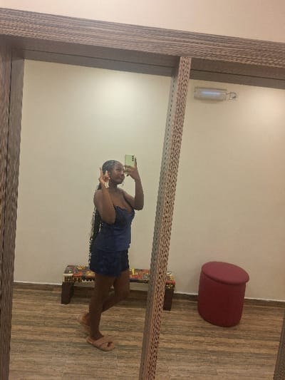 A mirror captures a woman in a blue dress, holding a phone, and making a peace sign. She stands on a wooden floor with a patterned frame. Behind her, a red ottoman sits near the wall, with books stacked beneath it. A cylindrical light fixture is mounted on the wall above. The mirror's frame has a striated, wood-like texture.