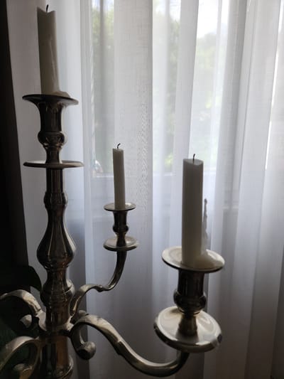 The image depicts an unlit, three-branched metallic candelabra, likely brass or a gold-toned alloy, positioned indoors in the city of Idar-Oberstein, Germany. Three white, cylindrical pillar candles are seated in the candelabra's holders. The candle on the far left is the tallest, appearing largely unburnt. The middle candle is significantly shorter, while the candle on the right is of medium height and exhibits visible wax drips along its side, indicating prior burning. All three candles feature exposed black wicks and are currently unlit. The candelabra itself is highly reflective, showing highlights and shadows across its ornate, polished surface, which features a central stem and three curved arms. In the background, a large window is obscured by multiple vertical panels of white, sheer, translucent curtains. Diffused natural light permeates through these curtains, revealing indistinct green foliage, likely trees or bushes, outside the window, suggesting a daytime scene. The overall lighting creates a backlight effect, highlighting the translucency of the curtains and casting the candelabra in partial silhouette with strong reflections. No individuals are present in the scene.