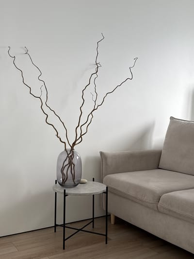 This is an interior scene located in Башкултаево, Russia. The image displays a light beige fabric sofa, partially visible on the right side of the frame, resting on light brown wooden floorboards. To the immediate left of the sofa, a small round side table with a light grey, concrete-textured top and black metal legs is positioned. On this table, a large, ovular, grey-tinted glass vase holds several tall, dark brown, gnarled bare branches, which extend vertically against a plain white wall, casting distinct shadows. A single small, light-colored, irregular stone rests on the table surface adjacent to the vase. No individuals or active subjects are present.