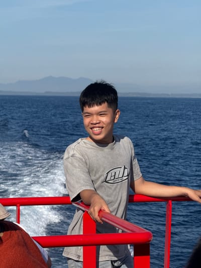 A young man with short black hair and a grey t-shirt stands on a red railing overlooking a body of blue water. The water behind him has a wake, indicating he is on a moving boat. In the distance, hazy mountains are visible under a clear blue sky. There are other people partially visible in the foreground on the left side of the frame. The young man is smiling and looking off to his right. The scene appears to be a daytime photograph taken on a ferry or boat. This specific location in Dong Son, Vietnam is characterized by its coastal geography and clear blue skies, as seen in the image.