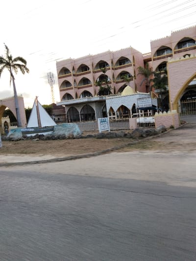 The image depicts an exterior view of a multi-story building complex in Gombato Bongwe ward, Kenya, observed from across an asphalt road under a bright sky. The prominent structure is a light peach-colored building, at least four stories high, characterized by a facade featuring multiple arched balconies or windows on each level and a decorative parapet along the roofline. Directly in front of this main building is a lower, single-story section with additional arches and a small golden dome, from which a sign advertising "FOR RENT APARTMENTS" with a phone number is visible. The complex perimeter includes decorative arched entrances to the left and right, and a low fence separates the property from the roadside.

In the foreground, an asphalt road runs horizontally across the lower portion of the frame, showing minor surface wear. Beyond the road, an unpaved median strip of dry, light brown earth with sparse vegetation extends towards the building. Positioned on this median is a white sailboat sculpture with a dark hull and two white sails, set upon a base of artificial rock formations painted in shades of light blue-green and brown. A smaller white sign with blue text is also present near the sculpture.

Vegetation includes several tall palm trees, one positioned to the far left and others within the complex grounds. In the background, a white lattice telecommunication tower rises to the left of the main building. The sky is largely clear and bright, indicating daylight conditions. No individuals are visible in the scene, and no specific actions or interactions are depicted.