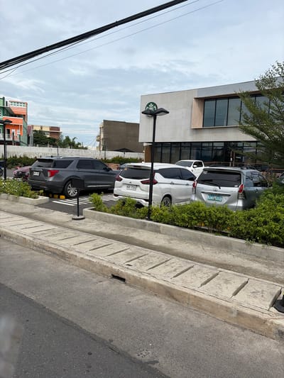 The image depicts a parking lot in front of a Starbucks coffee shop, located in Dagupan City, Philippines. Several cars are parked in the lot, including a grey Ford Explorer, a white Mitsubishi Xpander, and a silver Toyota Avanza. A Starbucks logo is visible on the side of the building, and a street lamp stands in front of the entrance. The weather is overcast, with a cloudy sky. In the foreground, there is a concrete curb and a section of asphalt road. The surrounding environment includes buildings with colorful facades, trees, and streetlights. The scene appears to be a typical urban setting on a cloudy day.