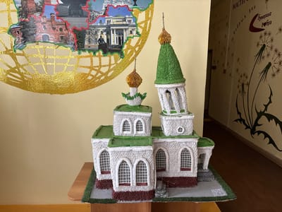 A model of a white church with a green roof is displayed on a wooden table. The church has multiple towers and arched windows with intricate white grids. The model sits on a green grassy base. Behind the church, a colorful collage is attached to a pale yellow wall. The collage depicts historical buildings and a statue within a red border, surrounded by a gold, textured grid pattern. To the right of the church, a hallway is visible with a dark brown floor and a pale yellow wall decorated with dark brown dandelion motifs and Cyrillic text. The overall scene appears to be an interior of a community center or educational institution.