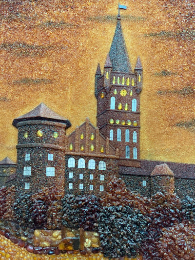 This media file displays a unique artwork, likely a mosaic or relief, crafted primarily from various shades and textures of amber and resin fragments. The scene depicts a stylized, multi-tiered cityscape or castle, with an implied medieval or fantastical aesthetic.

In the composition, a tall, slender spire dominates the left side, culminating in a simple weather vane. This spire and parts of the adjacent buildings are formed from fine, dark greyish-brown granular material. The background, filling the upper and left portions of the image, consists of lighter, golden-brown granular pieces, creating a textured surface that could represent a hazy sky or distant terrain, with faint vertical streaks of darker material.

The main architectural structures, occupying the center and right, are intricately detailed. Their facades are composed of a mosaic of small, irregularly shaped amber fragments, varying from light caramel to dark, rich brown, giving a rugged, textured appearance. Numerous windows are depicted: some are small, opaque white rectangles or squares, while others are represented by small, translucent, rounded yellow or orange amber pieces, suggesting illuminated interiors and lending an evening ambiance to the scene. One building prominently features a small, round yellow clock face with dark hands.

The base of the cityscape, particularly on the right, transitions into a dense accumulation of larger, irregularly shaped, dark amber pieces, resembling textured ground or foundations. Below this, a border of uniformly rounded, lighter amber pebbles forms a distinct edge. The artwork is devoid of any human figures or discernible text. The overall effect is a rich, tactile representation of an ancient or mythical settlement.