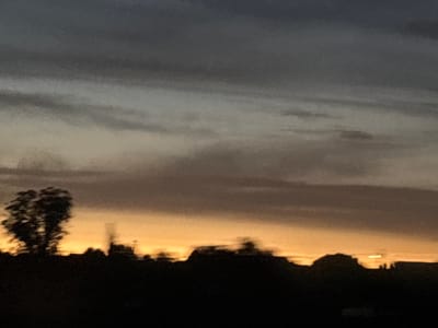 The image captures a silhouette of trees and a dark, undulating horizon against a dusky sky. The sky transitions from a deep gray at the top to a warm orange glow near the horizon, suggesting either sunrise or sunset. The silhouette in the foreground is composed of dark, indistinct shapes of trees and possibly low buildings. One prominent cluster of trees on the left is slightly more defined, with sparse branches. The overall impression is one of a rural or suburban landscape at twilight, with limited detail due to the low light and potential motion blur. There is no indication of specific locations within the city of Santarém, Portugal.