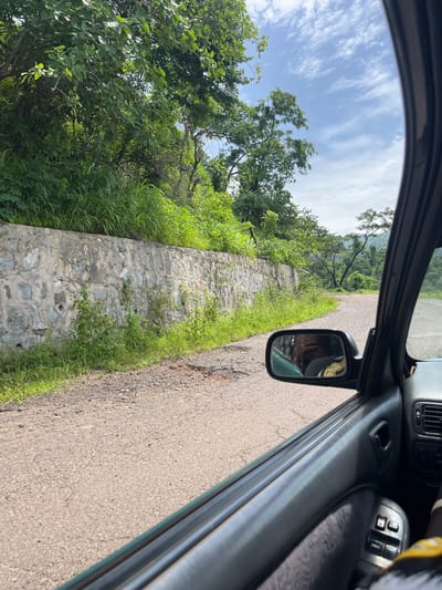 The image shows a view from inside a car, looking out the passenger side window. The car is on a paved road that appears to be in a rural or semi-rural area, with lush green vegetation and trees lining the left side of the road. A stone retaining wall is visible alongside the road, overgrown with grass and some small plants. The road curves to the right, leading into a landscape with rolling hills and more trees in the distance. The sky is partly cloudy. In the rearview mirror, a person can be seen, presumably the driver, wearing a yellow and black patterned shirt. The interior of the car, including the door panel, window controls, and part of the dashboard, is visible in the foreground. The scene suggests a journey through a scenic, natural environment, possibly on the outskirts of Goje, Nigeria, given the context.