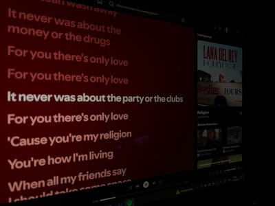 The image shows a screen display, likely a computer monitor, displaying the lyrics of a song by Lana Del Rey. The song appears to be "Religion" from the "Honeymoon" album. The lyrics are displayed in white text on a red background, with a portion of the playlist visible on the right side of the screen. The "Honeymoon" album cover is prominently featured. Additionally, the screen shows typical media player controls and other application functions at the bottom.