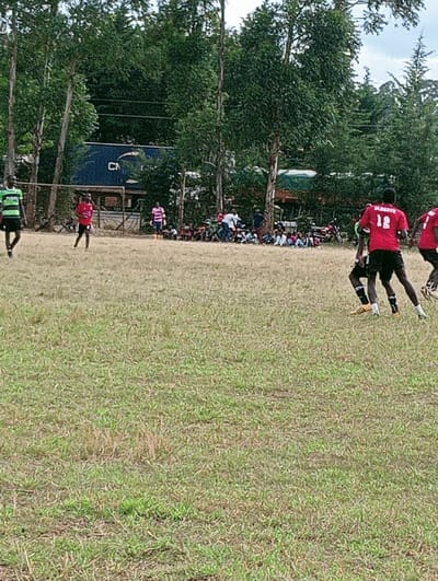 The scene depicts an outdoor football match or practice session in Lwandeti ward, Kenya. The foreground and mid-ground are dominated by a sparsely green grass field, showing areas of dry vegetation.
On the field, at least seven adult male individuals are visible, engaged in play. In the immediate right mid-ground, two players wear matching red jerseys with black shorts; one player wears jersey number "12" with "DAGOVE" printed on the back. Both are equipped with soccer cleats and appear in motion. To the left mid-ground, another player wears a green and black striped jersey with black shorts, moving away from the viewer. A fourth player in a red jersey and black shorts stands near a metal goal frame on the far left of the field. A fifth player in a pink and white striped jersey is positioned centrally in the mid-ground.
Along the far edge of the field, beneath a dense line of tall, leafy green trees, a group of approximately 20-30 spectators is seated or standing, observing the activity. This group includes both adults and children, with some children discernible in light-colored attire.
In the background, behind the trees and spectators, several objects are visible. A large blue shipping container, partially obscured by foliage, displays the letters "CN" on its side. Adjacent to it, a light-colored vehicle or truck is partly visible, alongside a green tarp-like structure. At least two motorcycles are parked among the spectators in the mid-ground background. The sky is overcast.